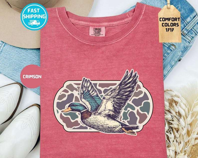 May include: Crimson-colored t-shirt featuring a detailed illustration of a duck in flight, set against a camouflage-style background. The shirt has a crew neck and short sleeves. The tag reads "Comfort Colors 1717".
