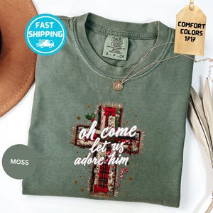 Puede incluir: Camiseta Comfort Colors verde musgo con un diseño de cruz. La cruz está decorada con cajas de regalo, bastones de caramelo y las palabras "oh come let us adore him". Un collar de oro está drapeado sobre la camisa.