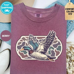 May include: Berry-colored sweatshirt featuring a detailed illustration of a flying duck with outstretched wings. The duck is set against a camouflage-style background within a rectangular frame. The shirt has a crew neck and the words "Comfort Colors 1717" are on a tag.