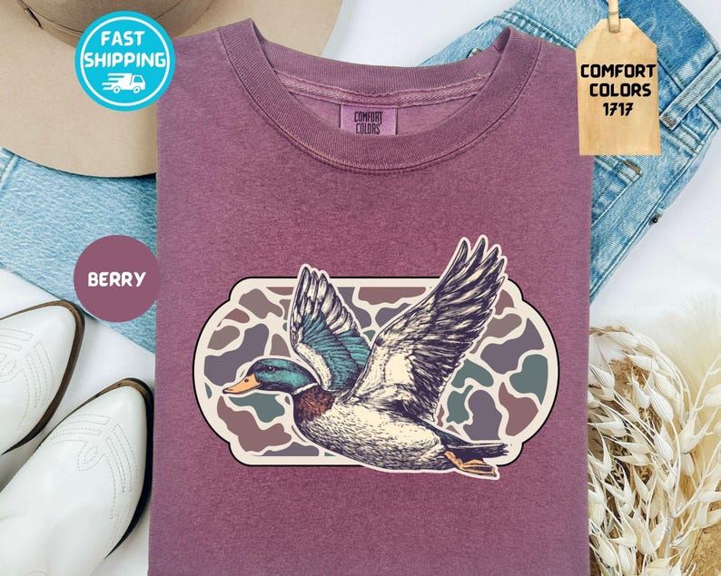 May include: Berry-colored sweatshirt featuring a detailed illustration of a flying duck with outstretched wings. The duck is set against a camouflage-style background within a rectangular frame. The shirt has a crew neck and the words "Comfort Colors 1717" are on a tag.