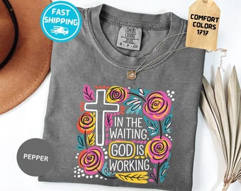 May include: A charcoal gray short-sleeve t-shirt featuring a graphic print of a white cross surrounded by stylized pink, yellow, and teal flowers. The text reads, IN THE WAITING, GOD IS WORKING. The shirt has a Comfort Colors 1717 label.