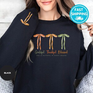 Pu&ograve; includere: Felpa nera con le parole "Grateful Thankful Blessed" in corsivo. La felpa presenta tre grafiche stilizzate a farfallino in arancione, giallo e verde. Il testo "GOD IS FAITHFUL IN EVERY SEASON" &egrave; in basso.