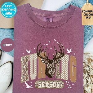 May include: Berry-coloured long-sleeve shirt with a deer head graphic and the words "HUNTING SEASON". The graphic includes a deer head, birds, and camouflage-patterned letters. The shirt is made of Comfort Colors 1717 material.
