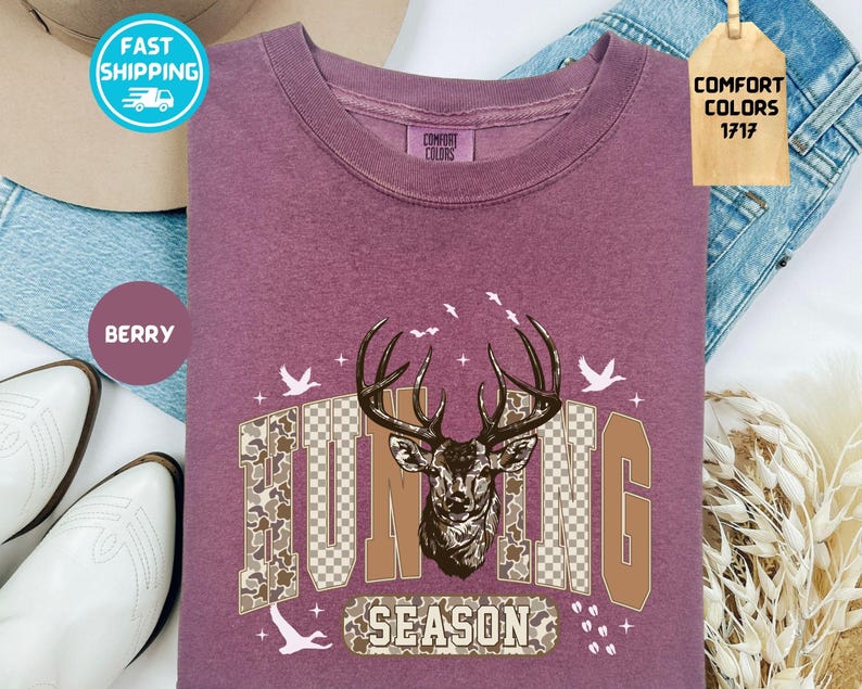 May include: Berry-coloured long-sleeve shirt with a deer head graphic and the words "HUNTING SEASON". The graphic includes a deer head, birds, and camouflage-patterned letters. The shirt is made of Comfort Colors 1717 material.