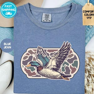 May include: Blue jean colored t-shirt with a graphic of a duck in flight. The duck is detailed with shades of blue, green, and brown. The shirt has a Comfort Colors tag and the text "BLUE JEAN".