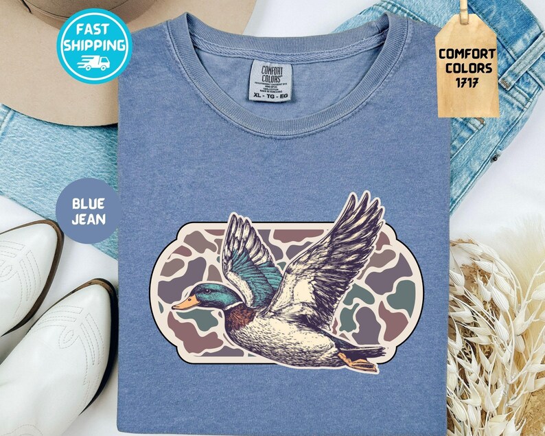 May include: Blue jean colored t-shirt with a graphic of a duck in flight. The duck is detailed with shades of blue, green, and brown. The shirt has a Comfort Colors tag and the text "BLUE JEAN".