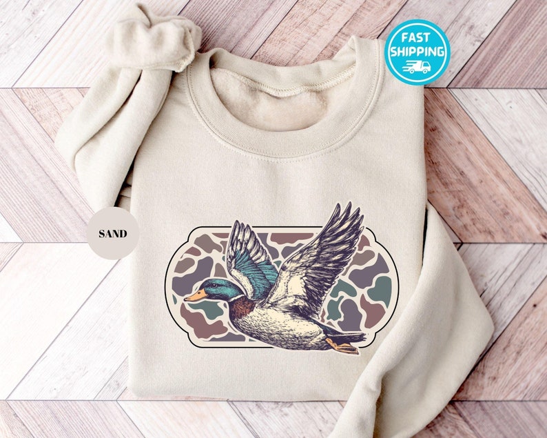 Peut inclure: Un sweat-shirt beige avec un imprim&eacute; graphique d'un canard volant au-dessus d'un motif camouflage. Le texte "SAND" est imprim&eacute; sur un cercle blanc.