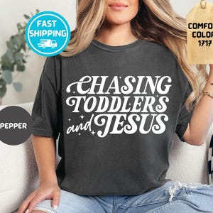 Op de afbeelding: Donkergrijs t-shirt met de witte tekst "CHASING TODDLERS and JESUS". Het shirt heeft een ronde hals en korte mouwen. Het woord "PEPPER" is gedrukt op een donkergrijze cirkel.
