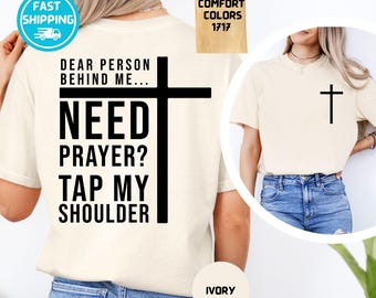 Puede incluir: Una camiseta color marfil con texto negro en la espalda que dice: DEAR PERSON BEHIND ME... NEED PRAYER? TAP MY SHOULDER, junto a una cruz. La parte delantera presenta una pequeña cruz negra en el pecho izquierdo.