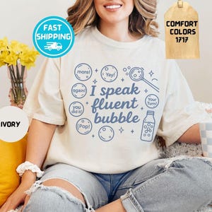 May include: Ivory-colored t-shirt with the phrase "I speak fluent bubble" in blue. The design includes bubble-themed graphics and text bubbles. The shirt has a relaxed fit and is part of the "Comfort Colors" collection.