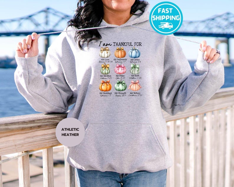 May include: Athletic heather gray hoodie with a graphic of colorful pumpkins and the text "I am THANKFUL FOR." The pumpkins are arranged in a grid, each with a different color and a corresponding word.