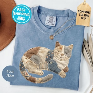 May include: A blue jean colored t-shirt with a patchwork cat design. The cat is made of various fabric patterns in shades of brown, blue, and cream. The shirt has a crew neck and short sleeves. The tag reads "Comfort Colors 1717".