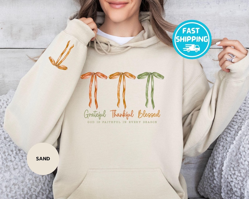 Pu&ograve; includere: Felpa con cappuccio color sabbia con cappuccio con coulisse e tasca frontale. Il design presenta fiocchi ad acquerello arancioni e verdi, con le parole "Grateful Thankful Blessed" stampate sotto. La manica ha un design a fiocco abbinato.