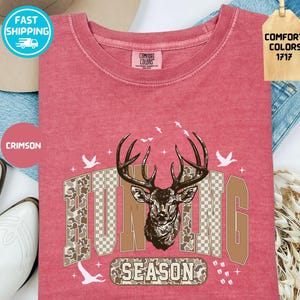 May include: Crimson t-shirt featuring a deer head graphic and the words "HUNTING SEASON." The design includes camouflage and a checkered pattern. The shirt has a crew neck and is made of Comfort Colors material.