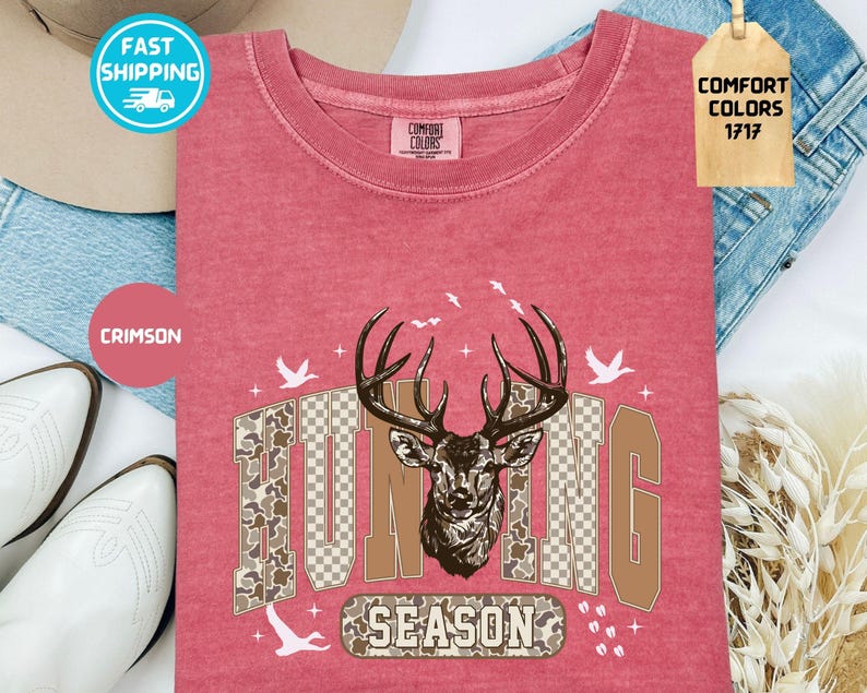 May include: Crimson t-shirt featuring a deer head graphic and the words "HUNTING SEASON." The design includes camouflage and a checkered pattern. The shirt has a crew neck and is made of Comfort Colors material.