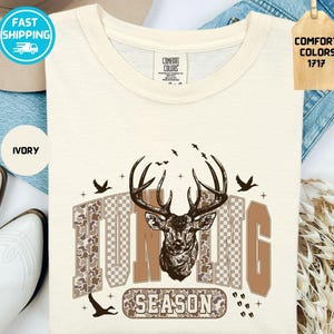 May include: Ivory t-shirt featuring a deer head graphic and the words "HUNTING SEASON". The design incorporates birds and a camouflage pattern. The shirt is from Comfort Colors.