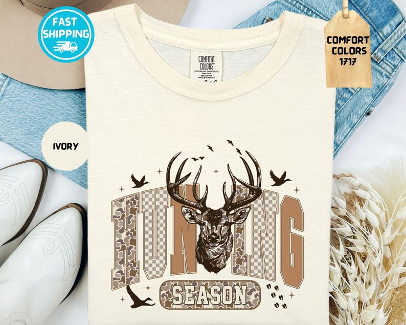 May include: Ivory t-shirt featuring a deer head graphic and the words "HUNTING SEASON". The design incorporates birds and a camouflage pattern. The shirt is from Comfort Colors.