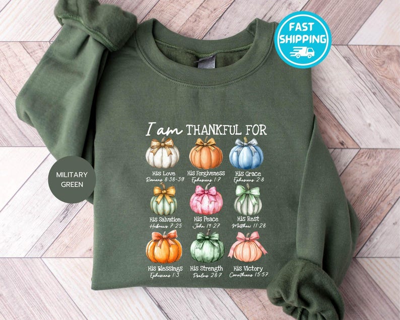 May include: Military green sweatshirt with the text "I am THANKFUL FOR" and colorful pumpkin illustrations. Each pumpkin represents a different blessing, with corresponding Bible verses. The sweatshirt has long sleeves and a crew neck.