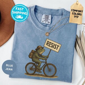 May include: Blue jean colored t-shirt with a frog riding a bicycle and holding a sign that says "RESIST". The frog is wearing a hat and backpack. The shirt has a Comfort Colors tag.