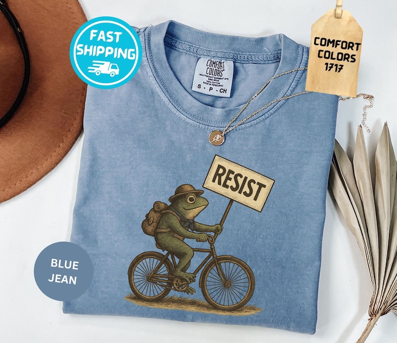 May include: Blue jean colored t-shirt with a frog riding a bicycle and holding a sign that says "RESIST". The frog is wearing a hat and backpack. The shirt has a Comfort Colors tag.