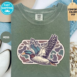 May include: A moss green t-shirt featuring a detailed illustration of a duck in flight, set against a camouflage-style background. The shirt has a classic crew neck and short sleeves. The tag reads "Comfort Colors 1717".