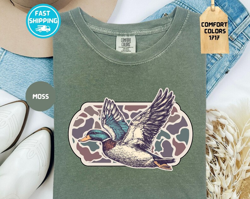 May include: A moss green t-shirt featuring a detailed illustration of a duck in flight, set against a camouflage-style background. The shirt has a classic crew neck and short sleeves. The tag reads "Comfort Colors 1717".