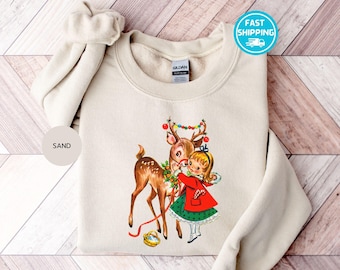 Op de afbeelding: Een crèmekleurige trui met een vintage illustratie van een meisje dat een rendier omhelst met een rode strik en een kerstverlichting. De tekst "SAND" staat op een beige cirkel in de linkerbenedenhoek van de afbeelding.