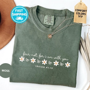 May include: A moss green short-sleeve t-shirt featuring the white text "fear not, for i am with you" and "ISAIAH 41:10" above a row of white daisies with orange centers and white butterfly outlines. The shirt has a Comfort Colors 1717 label.