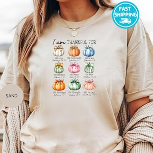 May include: Sand-colored t-shirt with the text "I am THANKFUL FOR" above nine watercolor pumpkins, each with a different color and a bow. Each pumpkin has a word and a Bible verse. A fast shipping sticker is in the top right.