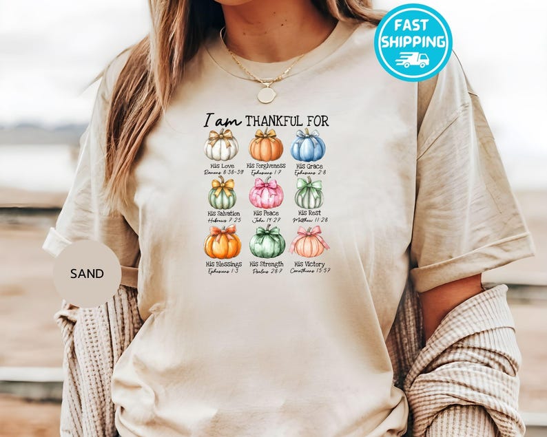 May include: Sand-colored t-shirt with the text "I am THANKFUL FOR" above nine watercolor pumpkins, each with a different color and a bow. Each pumpkin has a word and a Bible verse. A fast shipping sticker is in the top right.