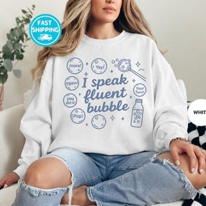 May include: White sweatshirt with the phrase "I speak fluent bubble" in blue lettering. The design includes bubble-related graphics and text like "more!", "Yay!", and "Pop!". The sweatshirt is a casual style.