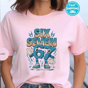 May include: A light pink t-shirt featuring the words "SIX SEVEN" in a blue and yellow font. Below the words is a cartoon-style number "67" with eyes, arms, and legs. The shirt has short sleeves and a round neckline.