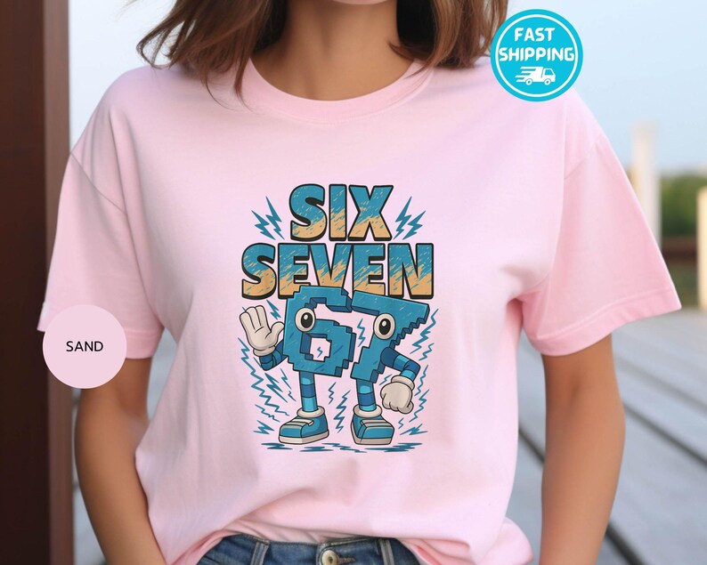 May include: A light pink t-shirt featuring the words "SIX SEVEN" in a blue and yellow font. Below the words is a cartoon-style number "67" with eyes, arms, and legs. The shirt has short sleeves and a round neckline.