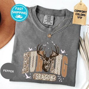 May include: A grey t-shirt featuring a deer graphic and the words "Hunting Season" in brown and camouflage. The shirt has a round neck and short sleeves. A tag reads "Comfort Colors 1717".
