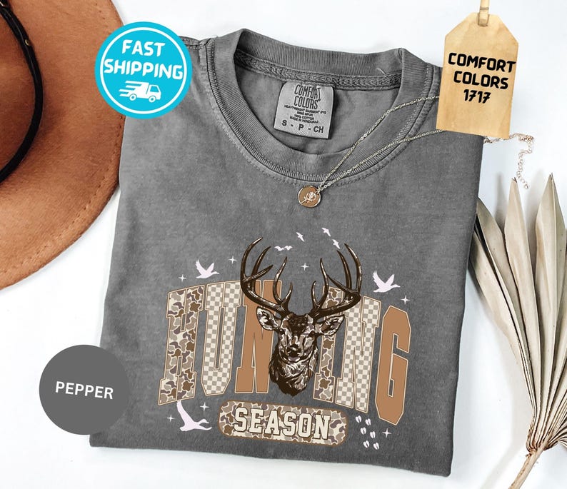 May include: A grey t-shirt featuring a deer graphic and the words "Hunting Season" in brown and camouflage. The shirt has a round neck and short sleeves. A tag reads "Comfort Colors 1717".