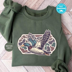 Peut inclure: Un sweat-shirt vert avec un motif camouflage et un dessin de canard en vol. Le texte "Military Green" est sur une &eacute;tiquette attach&eacute;e au sweat-shirt.