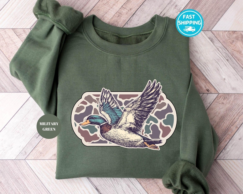 Peut inclure: Un sweat-shirt vert avec un motif camouflage et un dessin de canard en vol. Le texte "Military Green" est sur une &eacute;tiquette attach&eacute;e au sweat-shirt.