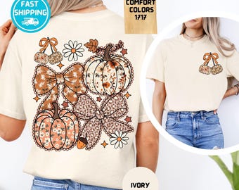 May include: An ivory t-shirt featuring a large back graphic of two pumpkins, two large bows, daisies, and small stars. The front left chest area displays a smaller graphic of two cherries with a bow. The text Comfort Colors 1717 is visible.