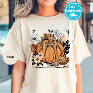 May include: Sand-colored t-shirt featuring a large, detailed pumpkin graphic with a plaid bow, surrounded by flowers and leaves. The design is framed by a black border. The word "SAND" is printed on a small circle.