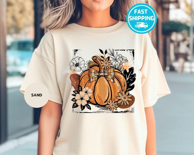May include: Sand-colored t-shirt featuring a large, detailed pumpkin graphic with a plaid bow, surrounded by flowers and leaves. The design is framed by a black border. The word "SAND" is printed on a small circle.