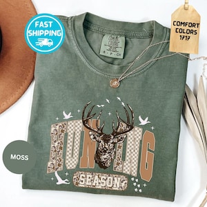 May include: A moss green t-shirt with a deer graphic and the words "HUNTING SEASON". The graphic features a deer head with antlers, camouflage and brown lettering. The shirt has a Comfort Colors tag.