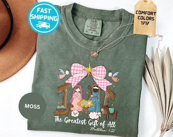 以下が含まれることがあります： モスグリーンのクルーネックTシャツ。キリスト降誕のシーン、ピンクのギンガムチェックのリボン、ラクダ、ロバ、羊のグラフィックが描かれています。テキストは「The Greatest Gift of All, Matthew 1:21」と書かれています。Comfort Colors 1717のラベルと小さなネックレスが付いています。