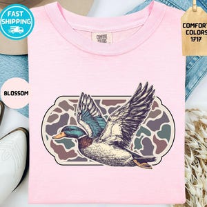May include: A light pink t-shirt with a graphic of a flying duck. The duck has detailed feathers and is set against a camouflage-style background. The shirt also has the text "Comfort Colors 1717" on a tag.