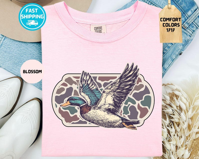May include: A light pink t-shirt with a graphic of a flying duck. The duck has detailed feathers and is set against a camouflage-style background. The shirt also has the text "Comfort Colors 1717" on a tag.