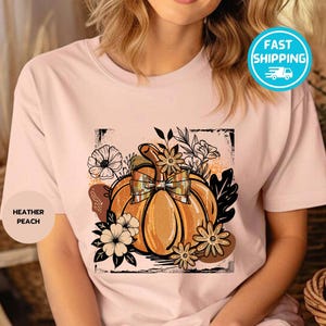 May include: Heather peach t-shirt featuring a pumpkin graphic with a plaid bow, surrounded by flowers and leaves. The design is in shades of orange, brown, and black, perfect for fall. The shirt has a round neck.