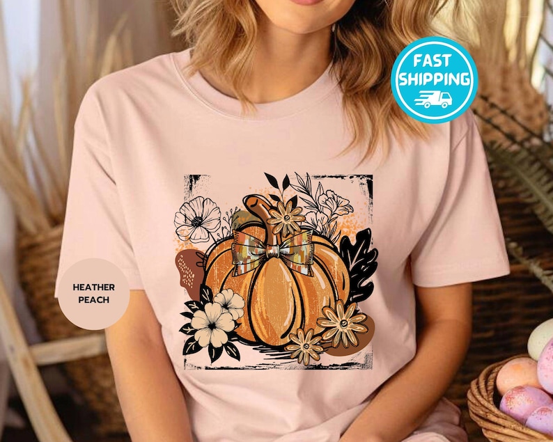 May include: Heather peach t-shirt featuring a pumpkin graphic with a plaid bow, surrounded by flowers and leaves. The design is in shades of orange, brown, and black, perfect for fall. The shirt has a round neck.