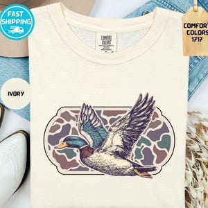 May include: Ivory-colored t-shirt featuring a detailed illustration of a duck in flight, with wings spread wide. The duck is set against a camouflage-style background within a decorative frame. The shirt has a classic crew neck and the words "COMFORT COLORS 1717" are visible.