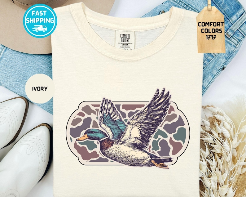 May include: Ivory-colored t-shirt featuring a detailed illustration of a duck in flight, with wings spread wide. The duck is set against a camouflage-style background within a decorative frame. The shirt has a classic crew neck and the words "COMFORT COLORS 1717" are visible.