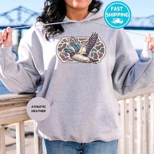 Peut inclure: Un sweat &agrave; capuche gris avec un imprim&eacute; de canard volant dans un motif camouflage. Le sweat &agrave; capuche porte les mots "Athletic Heather" imprim&eacute;s en bas &agrave; gauche.