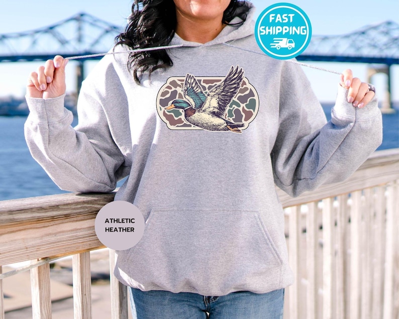 Peut inclure: Un sweat &agrave; capuche gris avec un imprim&eacute; de canard volant dans un motif camouflage. Le sweat &agrave; capuche porte les mots "Athletic Heather" imprim&eacute;s en bas &agrave; gauche.